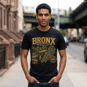 May include: Black t-shirt with a gold graphic that reads "BRONX Birthplace of Hip Hop Uptown NYC". The graphic includes an illustration of a bridge, buildings, and a map of the Bronx. The shirt is worn by a person standing on a city street.