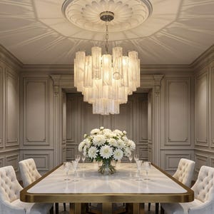 Op de afbeelding: Een meerlaagse kroonluchter met talloze verticale, doorschijnende witte glazen cilinders in een cirkelvormige opstelling. Het armatuur hangt aan een plafondrozet en verlicht een eetkamer met een wit bloemstuk als middelpunt.