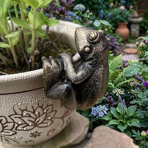 Puede incluir: Una escultura de rana de color bronce agarrada al borde de una maceta crema. La rana tiene una textura detallada y está colocada entre exuberantes plantas verdes y flores coloridas, creando una pieza decorativa con temática de jardín.