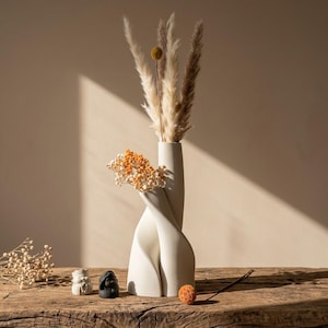 May include: A white, twisted ceramic vase displays dried flowers and pampas grass. Two small statues and an orange decorative ball are on a wooden surface. The vase has a unique, intertwined design, creating a modern, minimalist aesthetic.