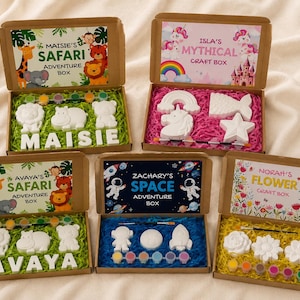 May include: Five open cardboard craft boxes containing white plaster figures, paint strips, and brushes. The boxes feature personalised labels: Maisie's Safari Adventure Box, Isla's Mythical Craft Box, Avaya's Safari Adventure Box, Zachary's Space Adventure Box, and Norah's Flower Craft Box.