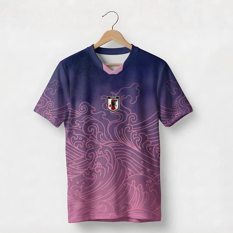 Op de afbeelding: Een marineblauw en paars shirt met korte mouwen en een roze golfpatroon. Het shirt heeft een embleem met het woord "JAPAN" en een rood-wit ontwerp. Het shirt hangt aan een houten hanger.
