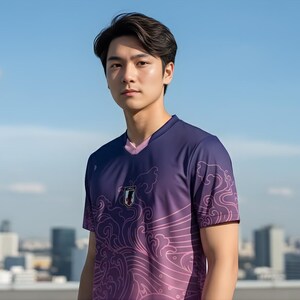 Op de afbeelding: Een paars en roze shirt met korte mouwen en een V-hals. Het shirt heeft een golfpatroon en het Japanse vlagembleem. Het shirt wordt gedragen door een persoon voor een blauwe lucht en een stadsgezicht.
