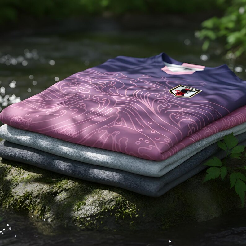 Op de afbeelding: Een stapel opgevouwen t-shirts, met een verloop van paars naar roze en een golfpatroon. Het bovenste shirt heeft een Japans vlagembleem. De shirts zijn gestapeld op een met mos bedekte rots bij water.