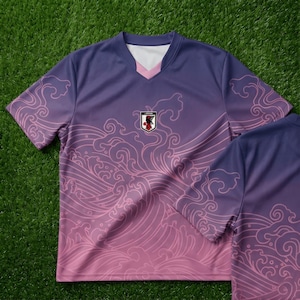 Op de afbeelding: Een paars en roze voetbalshirt met een golfpatroon. Het shirt heeft een V-hals met witte en roze afwerking en een embleem met de Japanse vlag. Het shirt met korte mouwen is gemaakt van lichtgewicht materiaal.
