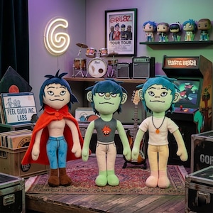 May include: Three plush dolls of animated band members from Gorillaz, each with unique outfits and hairstyles. The dolls stand on a patterned rug, with a drum set, arcade game, and band poster in the background. A neon "G" sign is also visible.