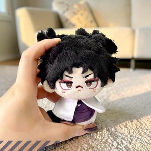 Puede incluir: Un muñeco de peluche con cabello negro y una expresión seria. El muñeco lleva un abrigo blanco sobre una prenda morada. El muñeco se sostiene en una mano, mostrando su pequeño tamaño. El muñeco tiene detalles rojos en los ojos y zapatos marrones.