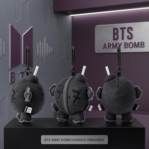 Può includere: Tre ornamenti in peluche grigio scuro a forma di bomba, ciascuno con una miccia bianca e una clip nera. Gli ornamenti hanno braccia e gambe piccole. Il testo "BTS ARMY BOMB" è visibile sullo sfondo. La parte inferiore del display recita "BTS ARMY BOMB HANGING ORNAMENT."