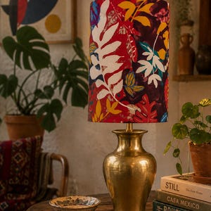 Könnte beinhalten: Eine Messing-Tischlampe mit einem farbenfrohen floralen Lampenschirm. Der Lampenschirm zeigt ein Muster aus weißen, roten, gelben und grünen Blättern. Die Lampe steht auf einem Holztisch mit Büchern und einer kleinen Schale. Ein dekorativer Einrichtungsgegenstand.