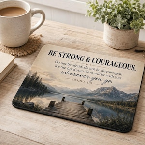 May include: A rectangular mousepad with the text "BE STRONG & COURAGEOUS" and a scenic landscape of a lake and mountains. A coffee mug, a book, and a small potted plant are also on the wooden surface.