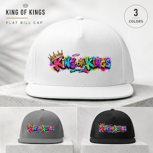 Op de afbeelding: Witte flat bill cap met de tekst "King of Kings" in kleurrijke graffiti-letters, een gouden kroon en een halo. De afbeelding toont de cap ook in grijs en zwart.