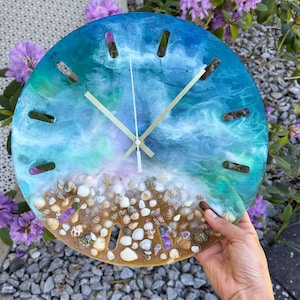 Könnte beinhalten: Eine runde Wanduhr mit Strandmotiv. Das Zifferblatt hat ein Harzdesign mit Blau- und Grüntönen, Muscheln und goldfarbenen Zeigern. Die Uhr wird vor einem Hintergrund aus lila Blumen gehalten. Durchmesser ca. 25 cm.
