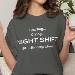 May include: Dark gray t-shirt with the text "Charting... Crying... NIGHT SHIFT Still Saving Lives" in white. The shirt is worn with denim shorts. A person is holding a white mug with a brown handle.