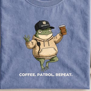 以下が含まれることがあります： ペリウィンクルブルーのTシャツ。タンカラーのパーカーと黒いセキュリティキャップを被り、コーヒーカップを持った緑色のカエルのグラフィックが描かれています。カエルはパーカーに無線機を付けています。下のテキストは「COFFEE. PATROL. REPEAT.」です。