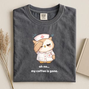 Op de afbeelding: Een antracietgrijs t-shirt met een afbeelding van een uil verkleed als verpleegkundige met een witte kap en uniform. De uil houdt een poot tegen het voorhoofd in een wanhopige houding. Onder de afbeelding staat: oh no... my coffee is gone.