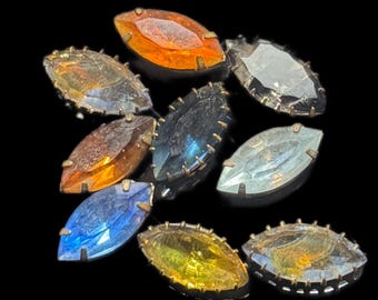 Könnte beinhalten: Eine Sammlung von marquiseförmigen Strasssteinen in verschiedenen Farben wie Orange, Blau, Gelb und Grau. Jeder Stein ist in einem goldfarbenen Metallrahmen gefasst, was einen Vintage-Look erzeugt. Die Steine sind vor schwarzem Hintergrund angeordnet.