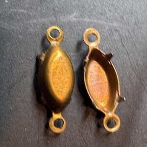 Könnte beinhalten: Zwei ovale Schmuckfassungen aus Messing mit goldfarbenem Metallic-Finish. Jede Fassung verfügt über einen vertieften ovalen Bereich in der Mitte mit vier kleinen Krallen sowie eine runde Öse an beiden Enden zur Befestigung an Ketten oder Elementen.