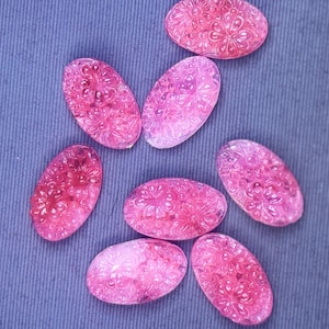 May include: Seven oval-shaped, translucent pink cabochons featuring a detailed, raised floral relief pattern across the surface. Each piece displays a consistent bright pink hue with a glossy finish and intricate carved flower petal details.