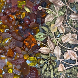 May include: A collection of amber-colored glass beads and faceted gems in various shapes and sizes, alongside metallic gold-toned filigree components featuring intricate leaf and floral patterns, arranged in a dense, mixed assortment.