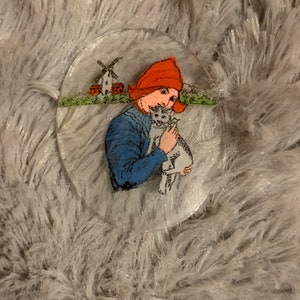 May include: A glass oval ornament with a painted image of a person wearing a red hat and holding a white animal. The person is standing in front of a windmill and a red building.