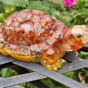 May include: A decorative turtle figurine made of clear resin with embedded orange and red elements. The turtle has a yellow base and feet, and is resting on a black metal surface. The background features green foliage and pink flowers.