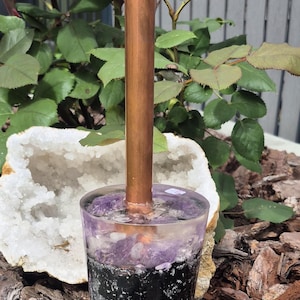Op de afbeelding: Een decoratief glazen object met een koperen buis die eruit oprijst. Het glas is gevuld met lagen amethist, zwarte steen en grijs grind. Het object staat tegen een achtergrond van groen en houtsnippers.