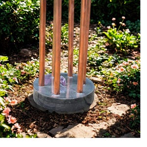 May include: A garden fountain made of copper and stone. The fountain has a dark grey stone base with several copper pipes rising from it, connected by copper wire. Amethyst crystals are visible inside the base. The fountain is in a garden setting.