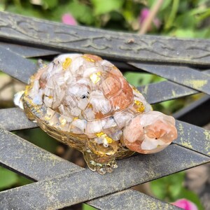 May include: A decorative turtle figurine with a clear, textured body and a shell made of clear and brown stones with gold flakes. The turtle is resting on a dark metal surface.