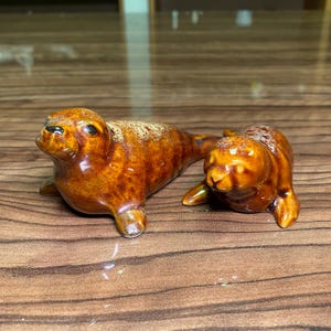 以下が含まれることがあります： 光沢のある茶色の釉薬が施された、2体の陶器製アザラシの置物。左側の大きいアザラシは正面を向き、右側の小さいアザラシは少しこちらを向いており、どちらも滑らかで光を反射する表面が特徴です。