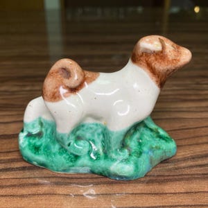 Może przedstawiać: Mała ceramiczna figurka psa z białym tułowiem oraz brązowymi łatami na głowie i ogonie. Pies stoi na teksturowanej, jaskrawozielonej podstawie imitującej trawę. Figurka posiada błyszczące wykończenie oraz zakręcony ogon.