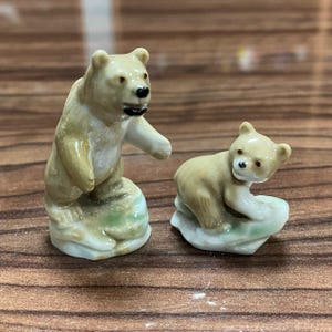 Könnte beinhalten: Zwei Miniatur-Bärenfiguren aus Keramik, eine aufrecht stehend und eine kleinere in kriechender Position. Beide Bären sind hellbraun mit weißen Akzenten, schwarzen Augen und schwarzen Nasen, jeweils auf einem kleinen, hellgrün-weißen Sockel.
