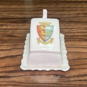 May include: A white porcelain butter dish with a rectangular base featuring a scalloped edge. The lid has a small handle on top and displays a colorful heraldic shield emblem with three lions, a crown, and a gold ribbon banner at the bottom.