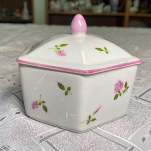 Puede incluir: Una caja de baratijas de porcelana blanca con tapa, con rosas rosas y hojas verdes. La tapa y la base están delineadas en rosa, y el remate también es rosa. La caja es cuadrada con base hexagonal.