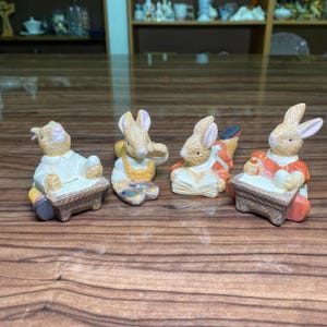 Peut inclure: Quatre figurines de lapins en céramique disposées en rangée. Le premier lapin est assis à un bureau, le deuxième tient une palette de peinture, le troisième est allongé en lisant un livre ouvert, et le quatrième est assis à un bureau avec un haut orange. Chaque lapin est peint en beige, blanc et diverses couleurs.