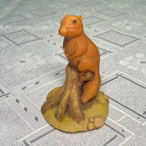 May include: A small, orange-brown figurine of a capybara perched atop a textured, light brown tree stump. The base of the sculpture is a rounded, yellowish-green mound with subtle rock-like details, creating a cohesive and detailed animal display.