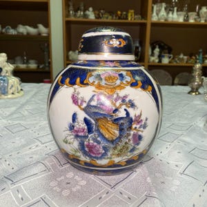 Peut inclure: Un pot en céramique blanc avec un couvercle, orné d'un motif de paon. Le pot a une forme ronde avec une bordure bleue et dorée. Le paon est représenté dans des tons de bleu, violet et or, entouré de fleurs roses.