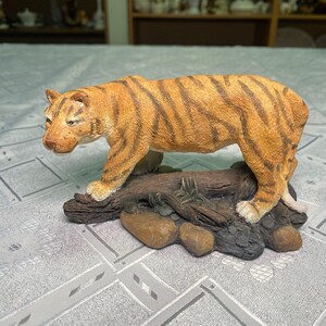 Op de afbeelding: Gedetailleerd beeldje van een tijger met oranje en zwarte strepen, geplaatst op een boomstam en rotsen. De tijger wordt afgebeeld in een loopstand, waarbij de realistische vachttextuur en gezichtskenmerken worden getoond. De basis is een mix van bruine en bruinachtige tinten.