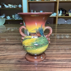 May include: A ceramic vase with two handles featuring a hand-painted landscape scene. The design depicts a house with a brown roof, green fields, and blue trees against a sunset-colored sky. The base and rim are a solid brown glaze.