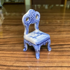 May include: A miniature porcelain chair featuring a white base with blue painted details. The backrest has an ornate lattice pattern, and the seat displays a blue landscape illustration of a windmill. The chair has four curved legs.