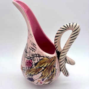 May include: A pink ceramic pitcher with a curved spout and a striped handle. The pitcher features a floral design with brown and yellow leaves, and pink and purple berries. The interior is a vibrant pink.