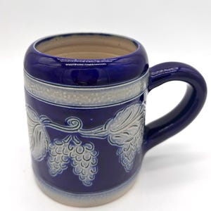 May include: A deep blue ceramic mug featuring a raised, light-colored relief design of hop cones and leaves wrapping around the exterior. The mug has a thick handle, a smooth glazed finish, and a light-colored interior rim and base.