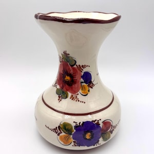 May include: A cream-colored ceramic vase with a scalloped rim and a brown painted edge. The body features hand-painted floral motifs in red, purple, yellow, and green, with a horizontal brown line encircling the midsection of the rounded base.