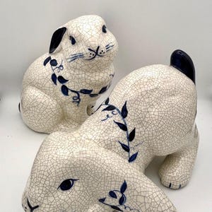 May include: Two ceramic rabbit figurines. The rabbits are white with a crackle glaze and dark blue accents. One rabbit is upright, and the other is lying down. The rabbits have dark blue ears and floral designs.
