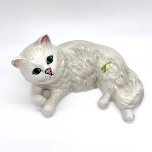 May include: A white ceramic cat figurine in a reclining pose. The cat has green eyes, pink ears, and a pink nose. The fur is textured, and a small green sticker is on its side. The figurine is on a white background.