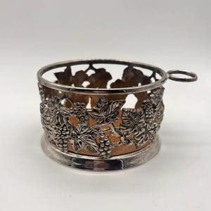 May include: A silver-toned metal coaster or glass holder featuring an intricate openwork design of grape clusters and vine leaves. The circular frame includes a small side handle and a textured, light brown cork-like base insert for stability.