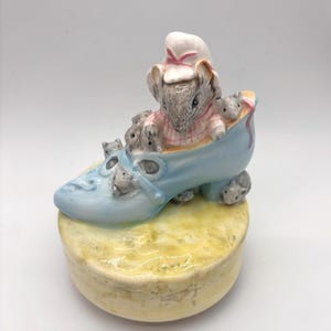 May include: A porcelain figurine featuring a mouse wearing a pink and white checkered shirt and a white bonnet, sitting inside a light blue shoe. Several smaller grey mice are sculpted onto the exterior of the shoe, which rests on a yellow base.