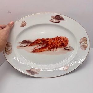 May include: An oval white porcelain serving platter featuring a central illustration of a bright orange lobster surrounded by various brown and tan seashells. The rim of the platter is finished with a thin metallic gold trim.