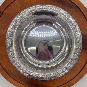 May include: A silver-colored bowl with a decorative floral pattern around the rim, set on a round wooden tray. The bowl's interior reflects the surroundings. A classic serving piece.