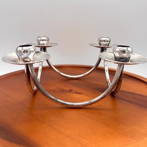 May include: A silver-toned metal candelabra featuring four circular candle holders connected by curved, arching supports. The structure is composed of polished metallic arms that form a symmetrical, interconnected base, designed to hold four individual candles.