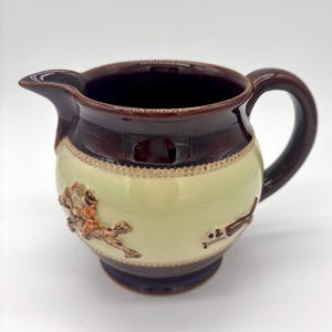 May include: A ceramic creamer pitcher featuring a two-tone design with a dark brown rim and base and a cream-colored center band. The center band is decorated with embossed figures of a person on horseback and a dog, framed by beaded borders.